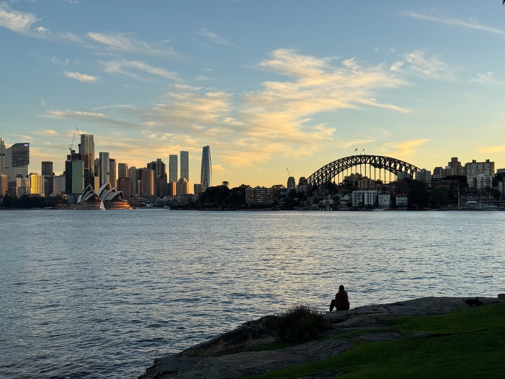 Image of a person at Cremorne Point sitting on a rock at sunset looking out over the harbour at the Opera House and Harbour Bridge 