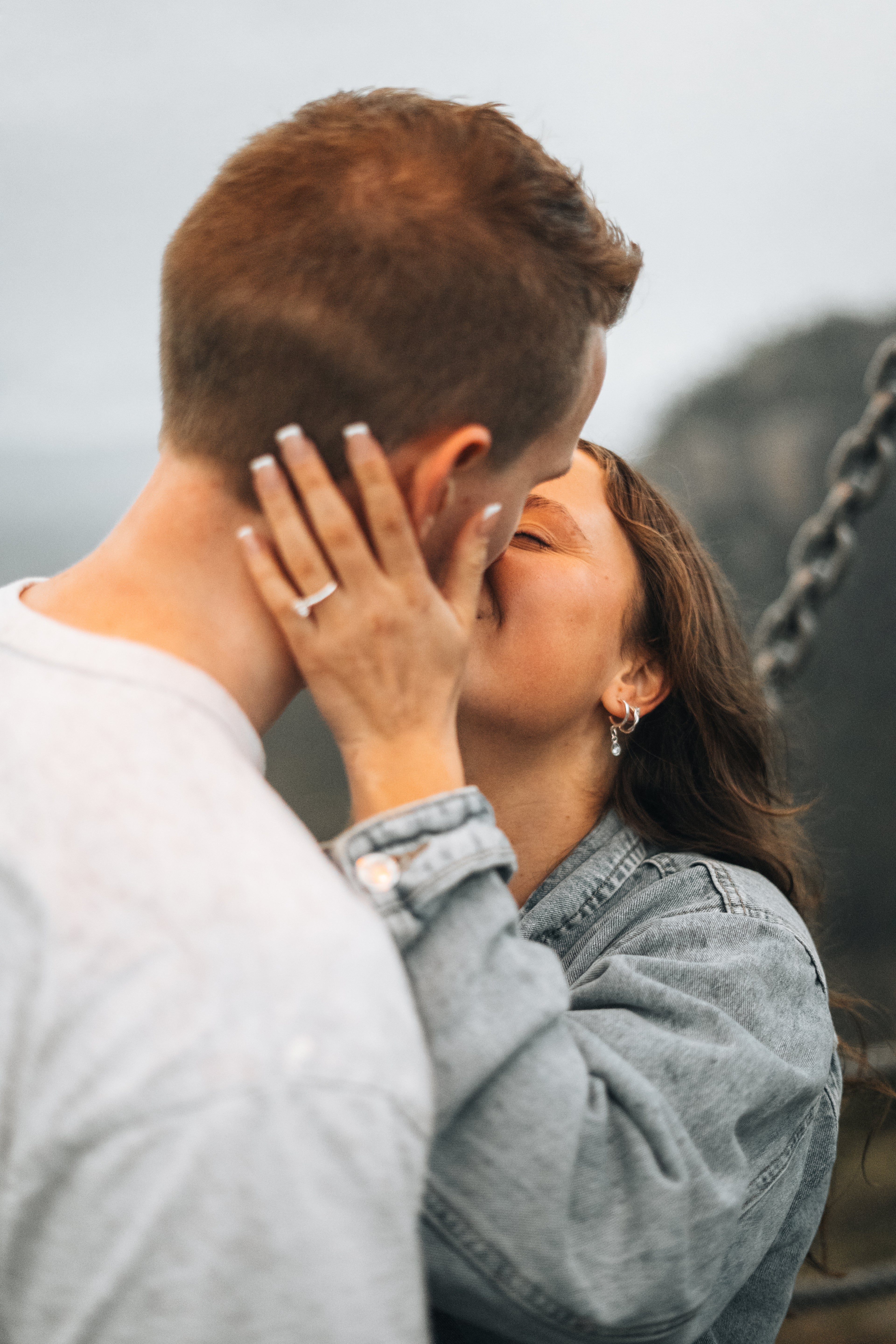 A couple embracing and kissing outdoors, with the woman wearing a light blue denim jacket and a silver ring on her finger, and the man in a light gray shirt. The background is a soft-focus natural setting with a chain visible on the right.