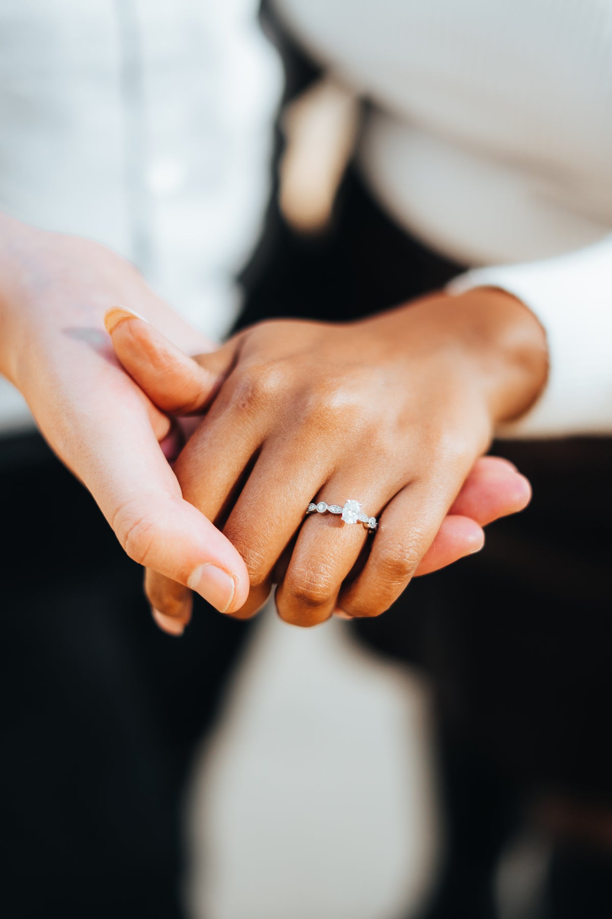 A close-up of two people holding hands, with a delicate silver band ring featuring a small clear gemstone worn on one finger. The background is softly blurred, showing white clothing.