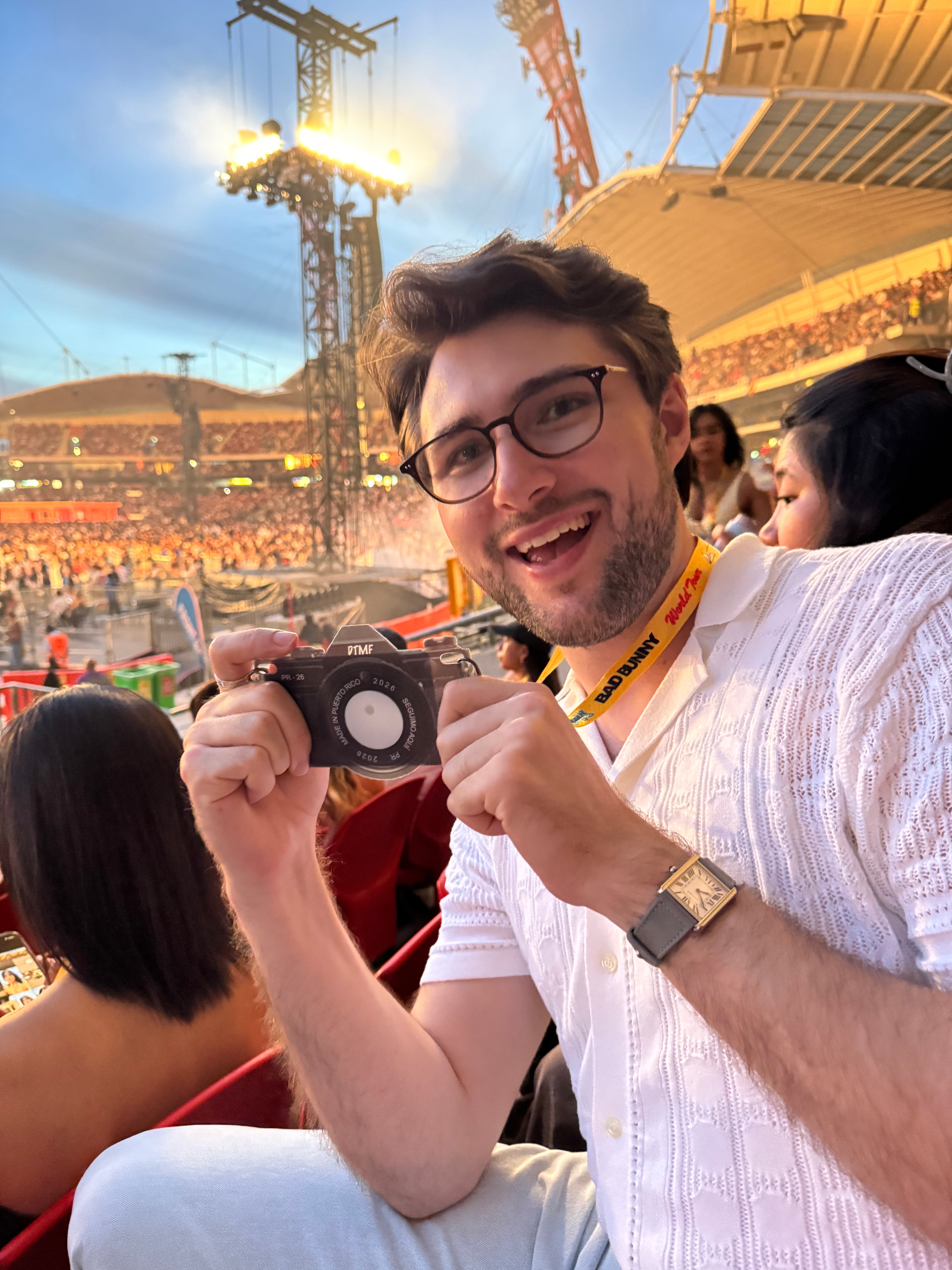 A man wearing glasses and a white textured shirt smiles while holding a black Nikon camera in a crowded stadium setting at sunset.